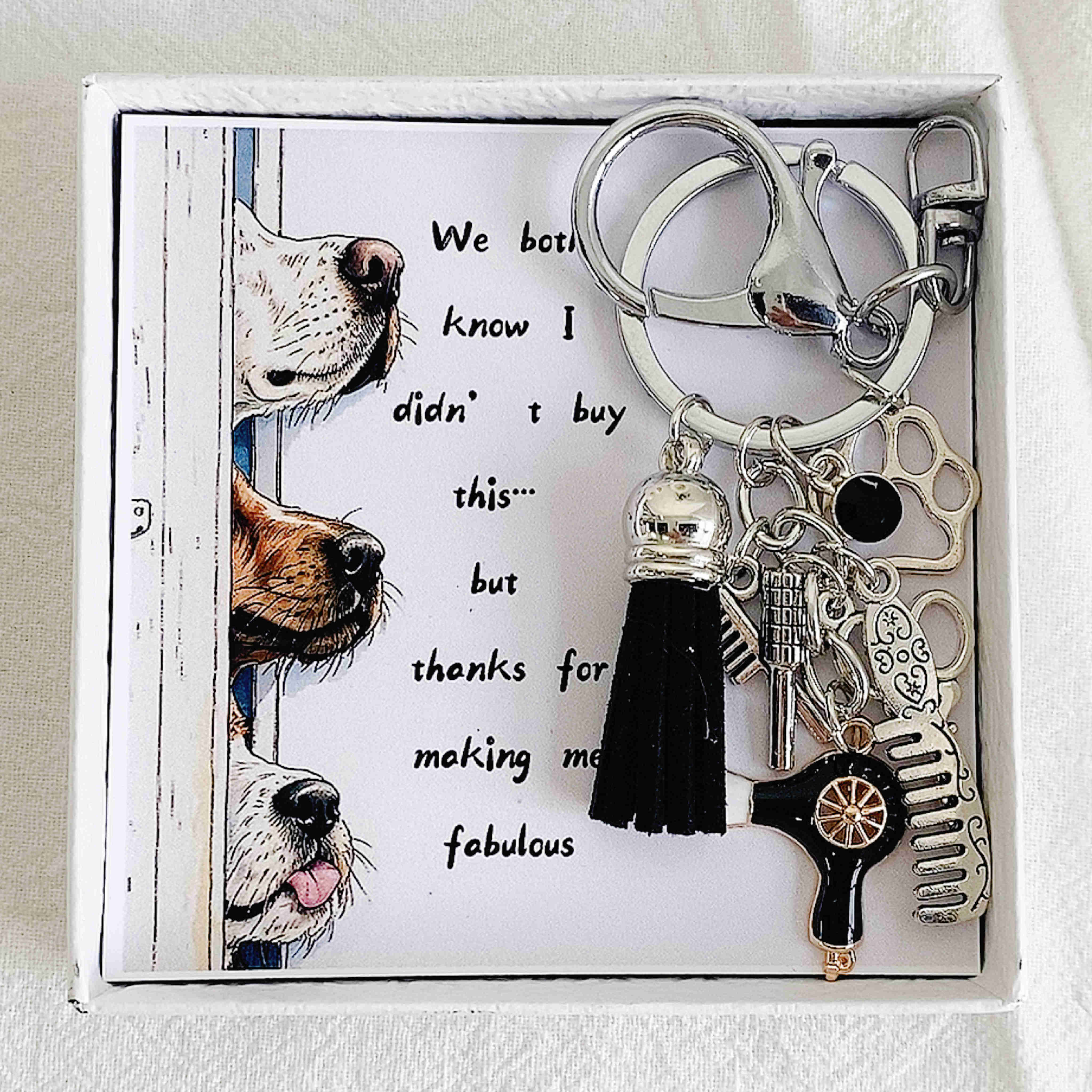 A handmade metal keychain with a tassel, designed with dog grooming tools such as hairdryer, comb, and scissors, presented in a box with a paw print design and text 'Good Hair Isn't By Accident, It's By Appointment'. The keychain also includes a cute paw print charm.