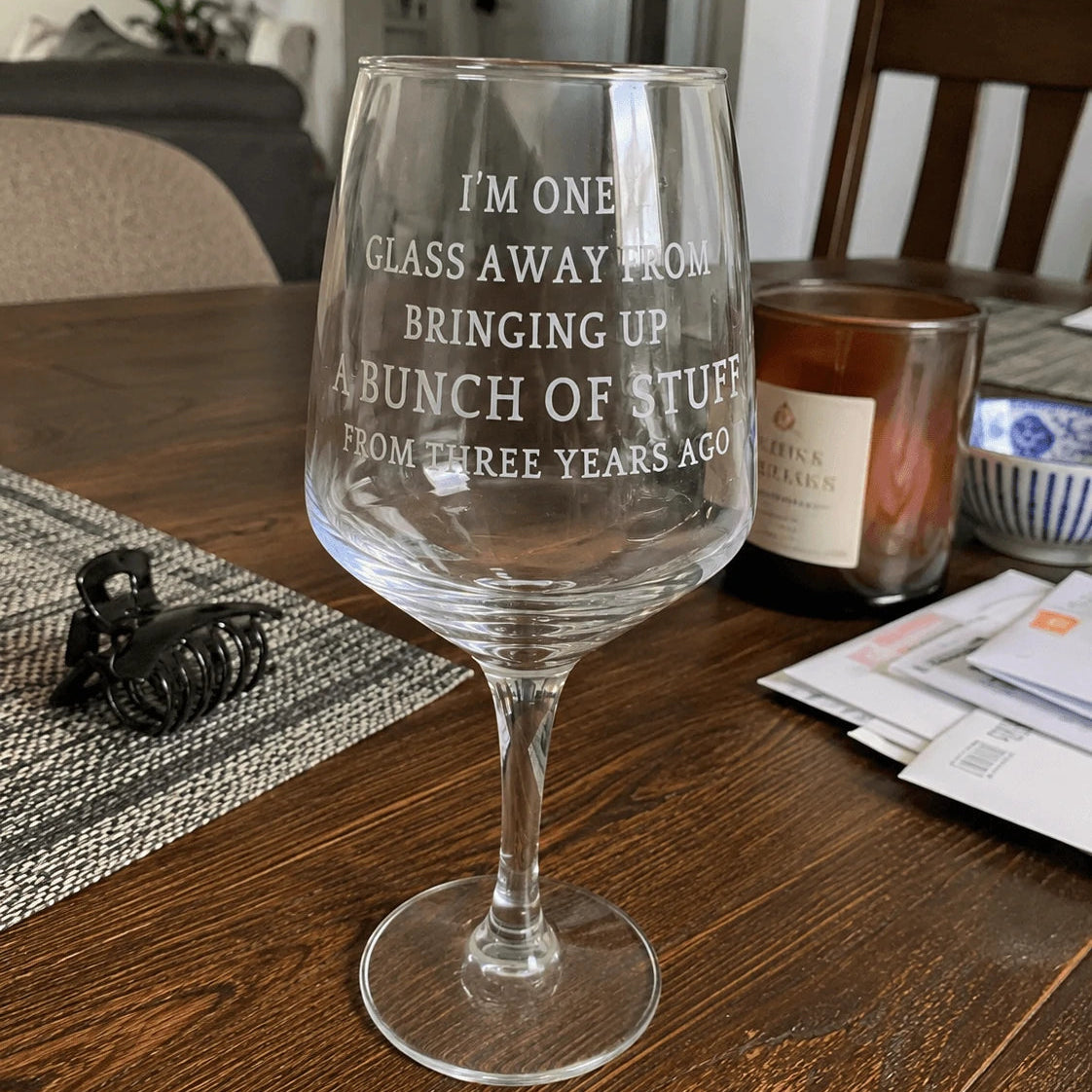 A clear wine glass with white frosted text reading 'I'M ONE GLASS AWAY FROM BRINGING UP A BUNCH OF STUFF FROM THREE YEARS AGO' sits on a wooden dining table in a home setting, surrounded by a candle, papers, and other household items.